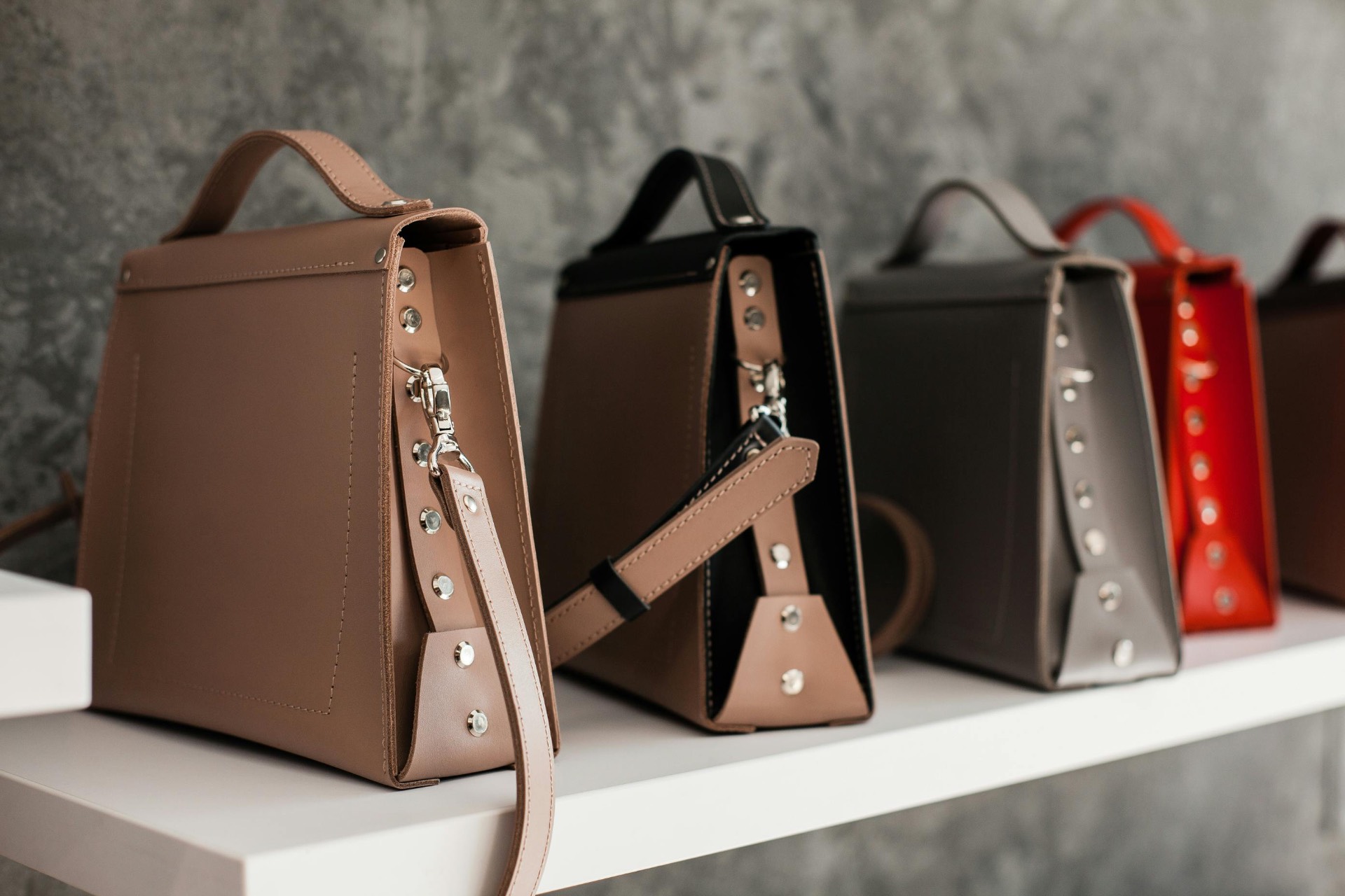 A row of handcrafted leather satchels in tan, black, grey and red on an atelier shelf